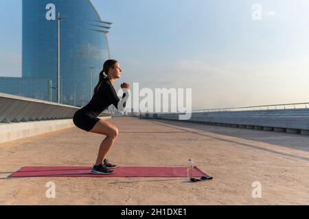 Vue latérale d'une athlète féminine en casque d'activité noir qui se claque les mains et fait des squats sur le tapis pendant l'entraînement physique tôt le matin en ville. Banque D'Images
