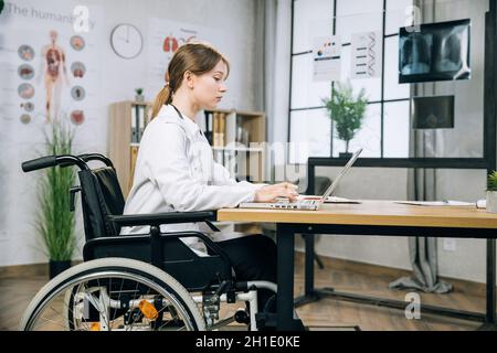 Femme de race blanche médecin utilisant le clavier en fauteuil roulant sur ordinateur portable au cabinet médical lumineux.Vue latérale d'un thérapeute qualifié sous une blouse de laboratoire travaillant sur un ordinateur portable à l'intérieur. Banque D'Images