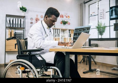 Spécialiste africain spécialisé dans le typage des personnes handicapées sur ordinateur portable, tout en analysant les résultats de l'examen radiographique.Spécialiste médical utilisant un fauteuil roulant travaillant dans une armoire moderne et lumineuse. Banque D'Images