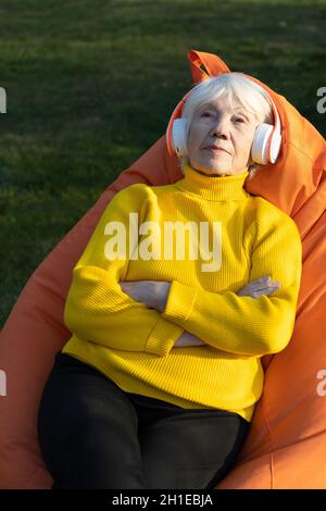 Belle femme de 85 ans aux cheveux gris, assise sur un fauteuil à sac et écoutant de la musique sur un casque. Banque D'Images