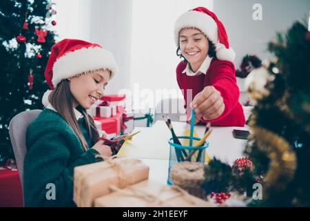 Photo des enfants gaies s'asseoir table préparer cadeaux écrire lettre de souhait porter x-mas chapeau pull dans la maison décorée à l'intérieur Banque D'Images