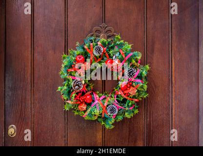 Vue de face d'une couronne de Noël colorée en branches de sapin en plastique et feuilles de houx avec des cônes et des rubans de pin, accrochée à un vieux doo de devant en bois Banque D'Images