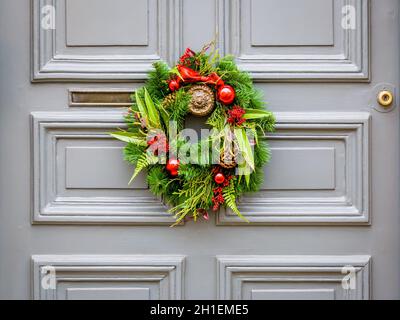 Vue avant d'une couronne de Noël en branches de sapin en plastique avec des cônes de pin, des boules et des rubans, accrochés à une porte avant grise avec moulures. Banque D'Images