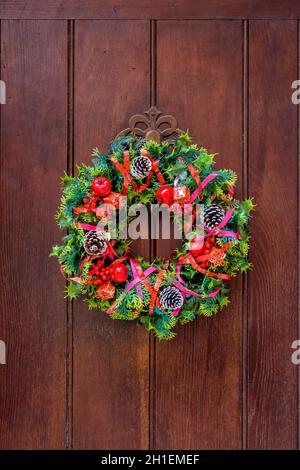 Vue de face d'une couronne de Noël colorée en branches de sapin en plastique et feuilles de houx avec des cônes et des rubans de pin, accrochée à un vieux doo de devant en bois Banque D'Images