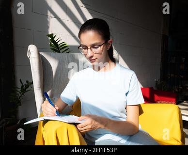 Belle jeune fille élève écrit avec un stylo une tâche dans un carnet tout en étant assis sur un fauteuil dans un café avec des ombres diagonales élégantes, lumière dure. Banque D'Images