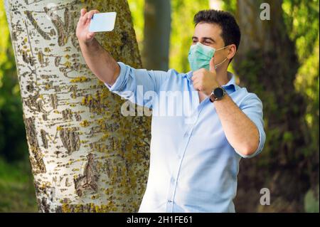 COVID-19 beau homme optimiste portant un masque chirurgical sur le visage pendant la maladie pandémique du coronavirus montrant les pouces dans le parc de la ville tout en faisant une vidéo Banque D'Images