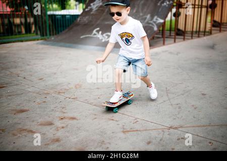 Garçon avec un skate dans un parc de skate. Un garçon avec des lunettes apprend à skate dans des vêtements élégants. Un peu extrême Banque D'Images