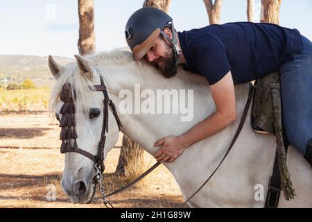 Un cavalier, un homme caucasien avec une barbe, est monté sur son cheval blanc et enserre son cou. Banque D'Images