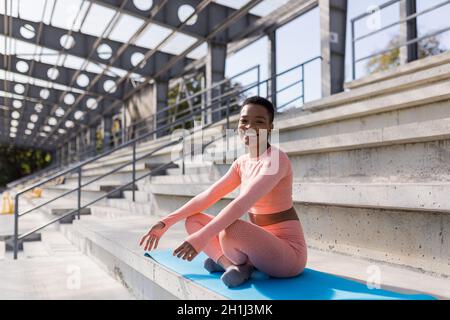 Femme afro-américaine souriante et regardant l'appareil photo, assise dans la position lotus sur le tapis de fitness, confiante et gaie après le yoga Banque D'Images