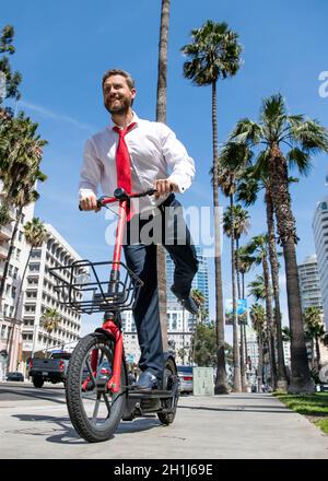 Un homme heureux en tenue de formalwear, e-scooter sur le trottoir, e-scoting Banque D'Images