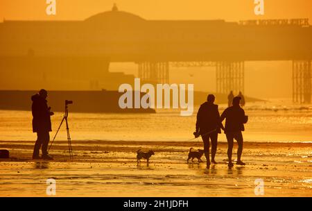 Plage de Brighton à marée basse, lever du soleil.Le Palace Pier a silhoueté en arrière-plan.Femmes marchant avec des chiens et photographe avec trépied.Brighton & Hove, Sussex, Angleterre, Royaume-Uni Banque D'Images