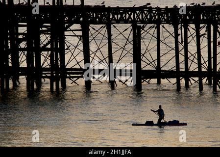 Pédalo en paddleboarder silhouetté autour de la jetée ouest abandonnée au coucher du soleil, Brighton, Angleterre.Construit en 1866 et fermé en 1975, le quai est toujours classé de catégorie I et un site bien connu. Banque D'Images