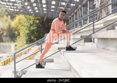 Athlète féminine en forme physique dans un costume de sport rose près du stade sportif, Afro-américain effectue des exercices de forme physique et des étirements le matin Banque D'Images