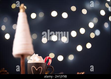 Une petite tasse de chocolat chaud surmontée de crème fouettée décorée de canne à sucre à côté d'une décoration de noël et de lumières de Noël floues en arrière-plan. Banque D'Images