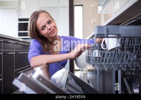 Jolie jeune femme dans sa cuisine équipée moderne et mettre dans le lave-vaisselle tasses - un appareil qui lui permet de garder la maison en ordre Banque D'Images