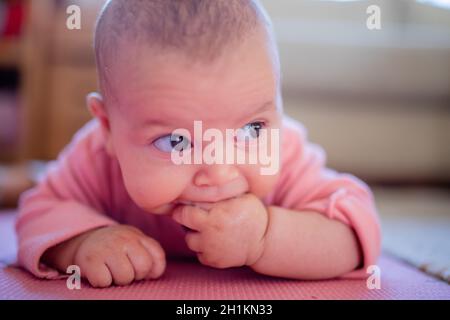 Bébé souriant dans une dors-bien rose couché face vers le bas sur un tapis rose avec deux de ses doigts dans la bouche Banque D'Images