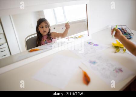 Paysage image d'un miroir sur une table blanche reflétant une petite fille dans une robe rose colorant différents types de feuilles sur une feuille de papier, avec Banque D'Images