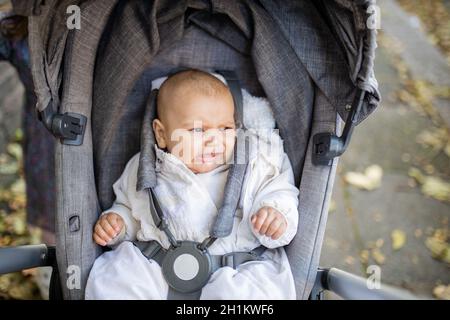 Photo d'un bébé en détresse dans une poussette grise regardant la distance et pleurant avec le trottoir couvert de feuilles d'automne comme arrière-plan Banque D'Images