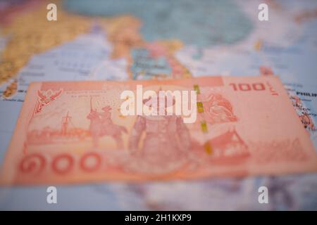 Gros plan de l'inverse d'un banknote de cent baht thaïlandais - avec le numéro 100 en thaï - en dessous de la Thaïlande sur une carte de l'Asie colorée et floue Banque D'Images