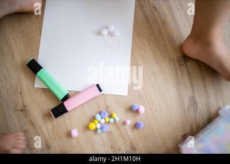 Photo de paysage d'une feuille de papier blanc, de marqueurs de texte et de boules de coton colorées sur un plancher en bois le long de la jambe d'un bébé et les jambes d'un littl Banque D'Images
