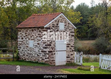 Magnifique springhouse en pierre restaurée avec toit rouge et porte et volets en bois blanc.Une clôture en bois blanc pittoresque se dresse à côté d'une porte au g Banque D'Images