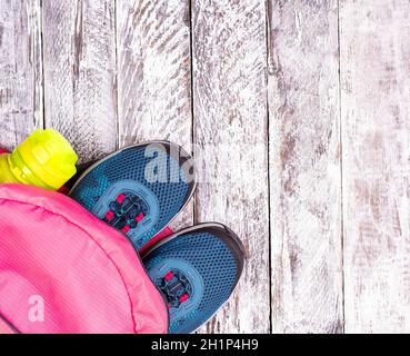 Paire de sneakers bleues pour femme dans un sac à dos rose et une bouteille de sport pour une eau sur une surface en bois blanc Banque D'Images