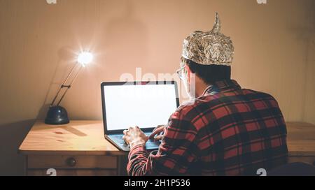 Un jeune homme avec un capuchon en aluminium est assis dans le sous-sol sombre devant un ordinateur portable. Concept de la théorie du complot Banque D'Images