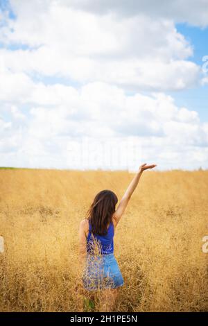 Portrait d'une jeune belle femme brune dans un t-shirt bleu et un short en denim se tient au milieu du champ, exposant son visage au soleil sur le Banque D'Images