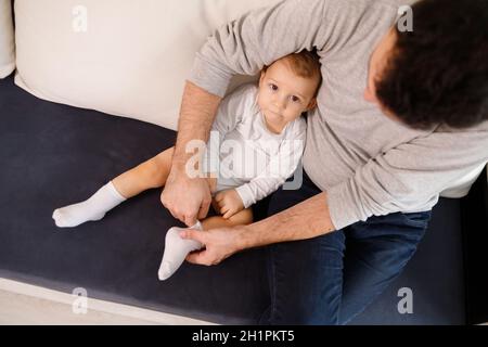 Le jeune père met des chaussettes sur son fils, assis sur un canapé. Banque D'Images