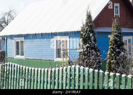 Ancienne maison en bois dans le village biélorusse.Maison en bois avec une clôture faite de planches avec de l'épinette verte en hiver. Banque D'Images