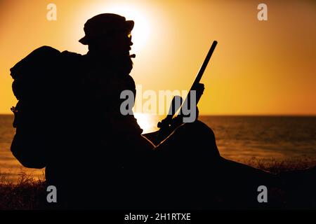 Silhouette de commando combattant, de tireur des forces spéciales de l'armée assis sur la mer ou sur la rive de l'océan au coucher du soleil.Garde côtière rifleman observant la plage, duri de repos Banque D'Images