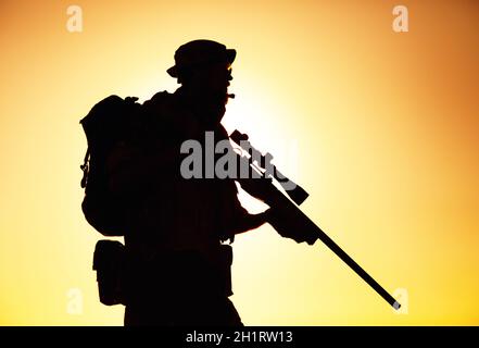 Silhouette de sniper des forces d'élite de l'armée, tireur de commando en chapeau de boonie, porte sac à dos, radio tactique équipée, marche sur fond de mise o Banque D'Images