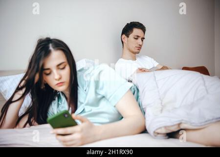 Couple assis dans le lit arguant et maintenant ils sont contrariés les uns avec les autres, la femme l'ayant retourné sur l'homme écrivant des messages sur le téléphone Banque D'Images