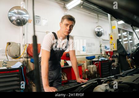 Portrait moyen d'un mécanicien de voiture de qualité professionnelle en uniforme bleu debout devant le capot ouvert. Banque D'Images