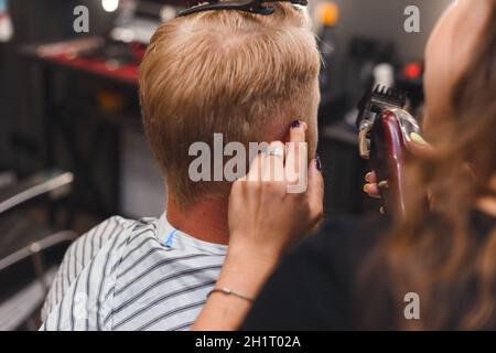 Le masque de coiffeur féminin coupe les poils d'un homme avec une tondeuse à cheveux. Coiffure pendant la prise de distance sociale. Banque D'Images