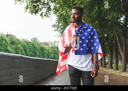 Beau Afro américain homme avec drapeau des Etats-Unis sur ses épaules regardant sérieusement la caméra, debout à l'extérieur. Jour d'été Banque D'Images