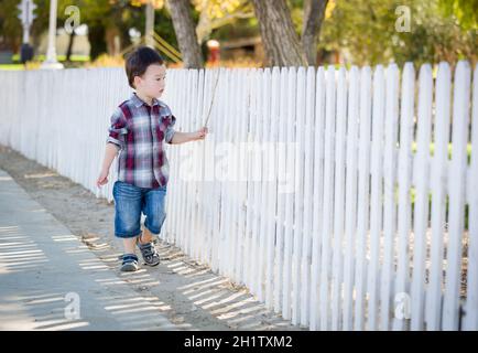 Cute Young Mixed Race Boy Walking Stick avec le long de clôture blanche. Banque D'Images