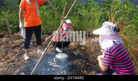 Groupe de jeunes filles asiatiques qui cuisent dans un pot noir autour d'un feu de camp dans une forêt extérieure. Banque D'Images