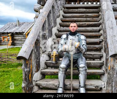 Chevalier dans l'armure sur le marches de bois. Armure chevaleresque et arme. - Antique semi photo. Banque D'Images