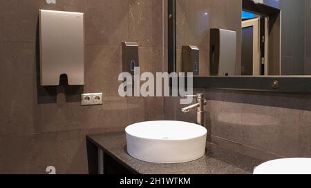 Robinets chromés avec lavabos ronds blancs dans une toilette publique avec un grand miroir et des murs gris, intérieur moderne d'une toilette publique Banque D'Images