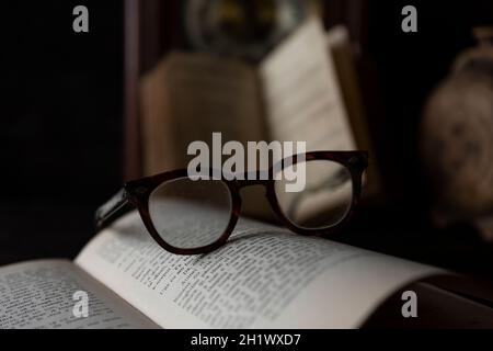 Vieilles lunettes usées reposant sur un livre ouvert avec des livres et une horloge en arrière-plan. Notion : temps de lecture Banque D'Images