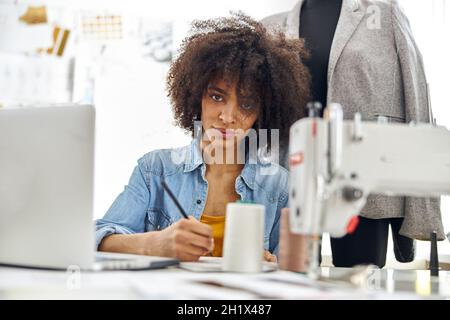 La jolie couturière afro-américaine s'assoit à table avec une machine à coudre moderne Banque D'Images