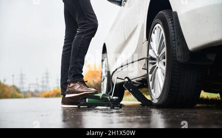 Gros plan des jambes de sexe masculin dans un Jean gris gonflant un pneu crevé avec un compresseur d'air.Jeune homme utilisant un compresseur d'air pour gonfler le pneu en se tenant près de la voiture sur route pluvieuse. Banque D'Images