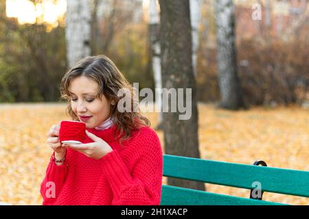 Une belle jeune femme dans un chandail et un bracelet rouge vif est assise sur un banc de parc et tient une tasse rouge avec une boisson chaude dans ses mains contre le dos Banque D'Images