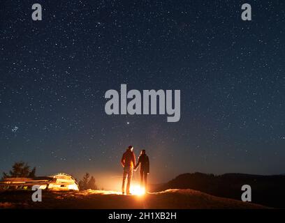 Vue arrière du couple en amour debout, tenant les mains, feu de camp à proximité et admirer l'environnement romantique dans le camp de tente contre le ciel étoilé magique.Concept de randonnée, camping de nuit, relations Banque D'Images