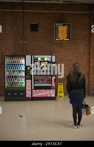 Doncaster, Royaume-Uni, 22 mai 2021 : une jeune femme attend patiemment près des distributeurs automatiques, tenant son sac à main, regardant le panneau de départ du train Banque D'Images
