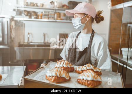 Une employée de masque médical tient des croissants fraîchement cuits dans la cuisine du magasin Banque D'Images