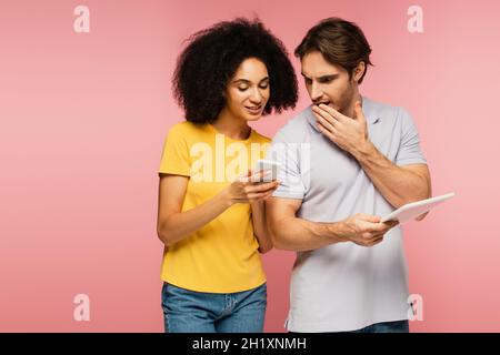 femme hispanique souriante montrant un smartphone à l'homme choqué avec la tablette numérique isolée sur rose Banque D'Images