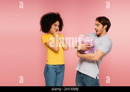 femme hispanique souriante touchant le visage tout en regardant l'homme stupéfié avec beaucoup de cadeaux isolés sur le rose Banque D'Images
