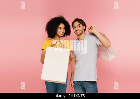 homme joyeux souriant à l'appareil photo près d'une femme hispanique avec sac de shopping isolé sur rose Banque D'Images